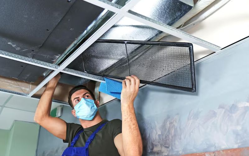 A masked technician in overalls cleaning a ceiling air vent grille below exposed ductwork